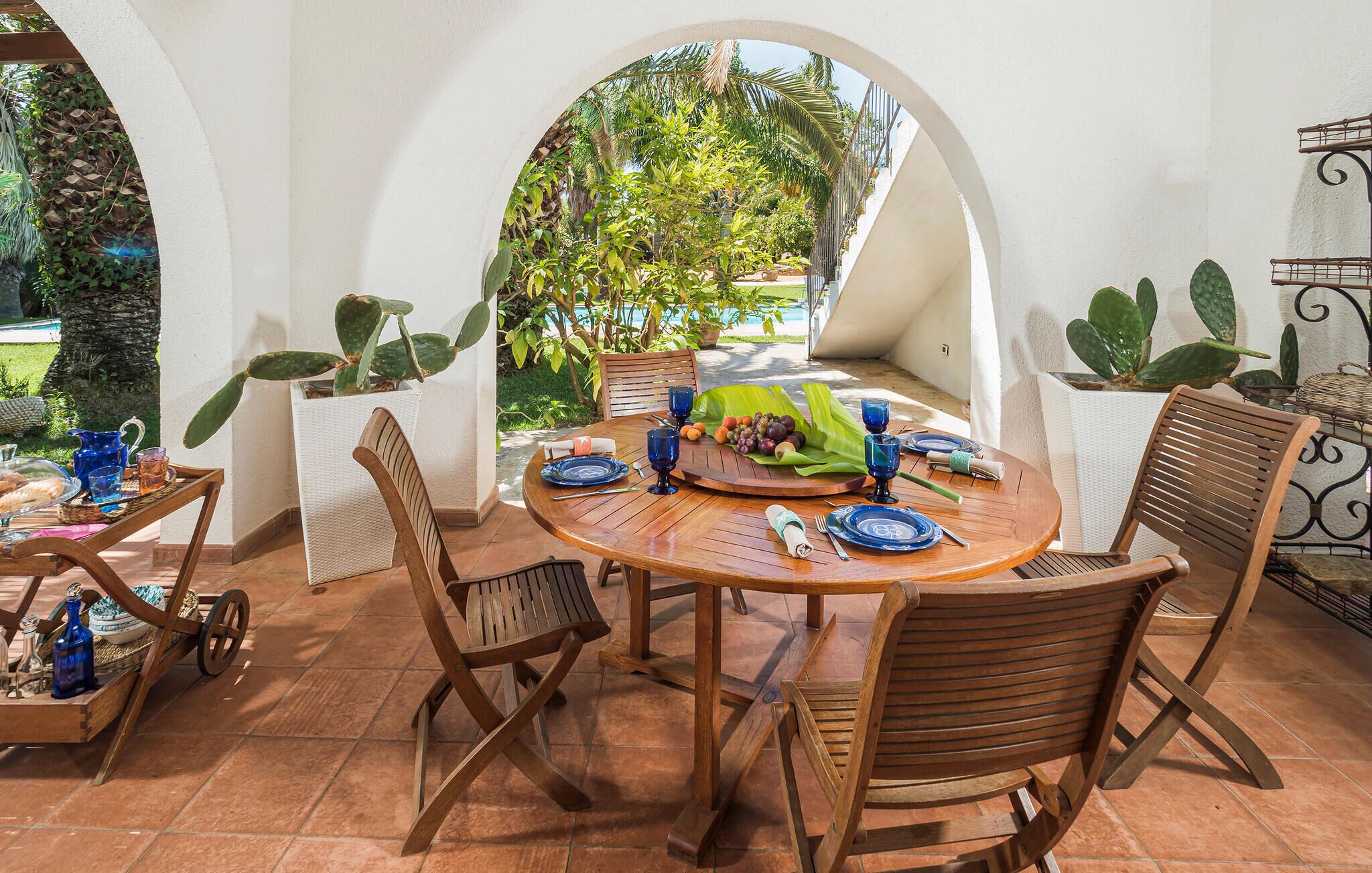 An outdoor dining area with a round wooden table, chairs, and a view of a pool and greenery.