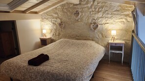 A bedroom with a large bed, two bedside tables with lamps, a stone wall with heart-shaped decorations, and a wooden floor.