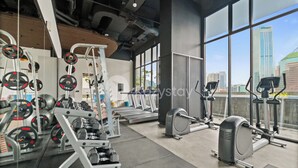 Un gimnasio con diverso equipamiento de ejercicio, ventanas amplias y vistas a la ciudad.