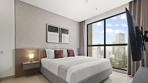Una habitación de hotel moderna con una cama grande, una mesita de noche con lámpara, vista a la ciudad a través de un ventanal amplio y un televisor montado.