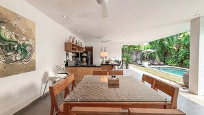 A dining area with a glass-top table and wooden chairs, a kitchen with wooden cabinets, and a view of a swimming pool and lush greenery. - Villa Alexia in Seminyak - 5 minutes walk to Beach (Seminyak)
