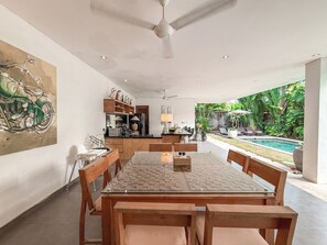 A dining area with a glass-top table and wooden chairs, a kitchen with wooden cabinets, and a view of a swimming pool and lush greenery. - Villa Alexia in Seminyak - 5 minutes walk to Beach (Seminyak)