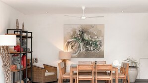 A dining area with a table, chairs, a ceiling fan, a wall-mounted painting of a motorcycle, and a shelving unit with decorative items. - Villa Alexia in Seminyak - 5 minutes walk to Beach (Seminyak)
