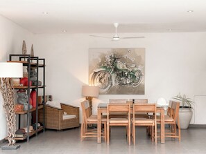A dining area with a table, chairs, a ceiling fan, a wall-mounted painting of a motorcycle, and a shelving unit with decorative items. - Villa Alexia in Seminyak - 5 minutes walk to Beach (Seminyak)