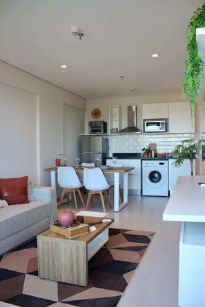 A modern living room with a sofa, dining table, chairs, and a coffee table. The kitchen area includes a microwave, oven, and washing machine.