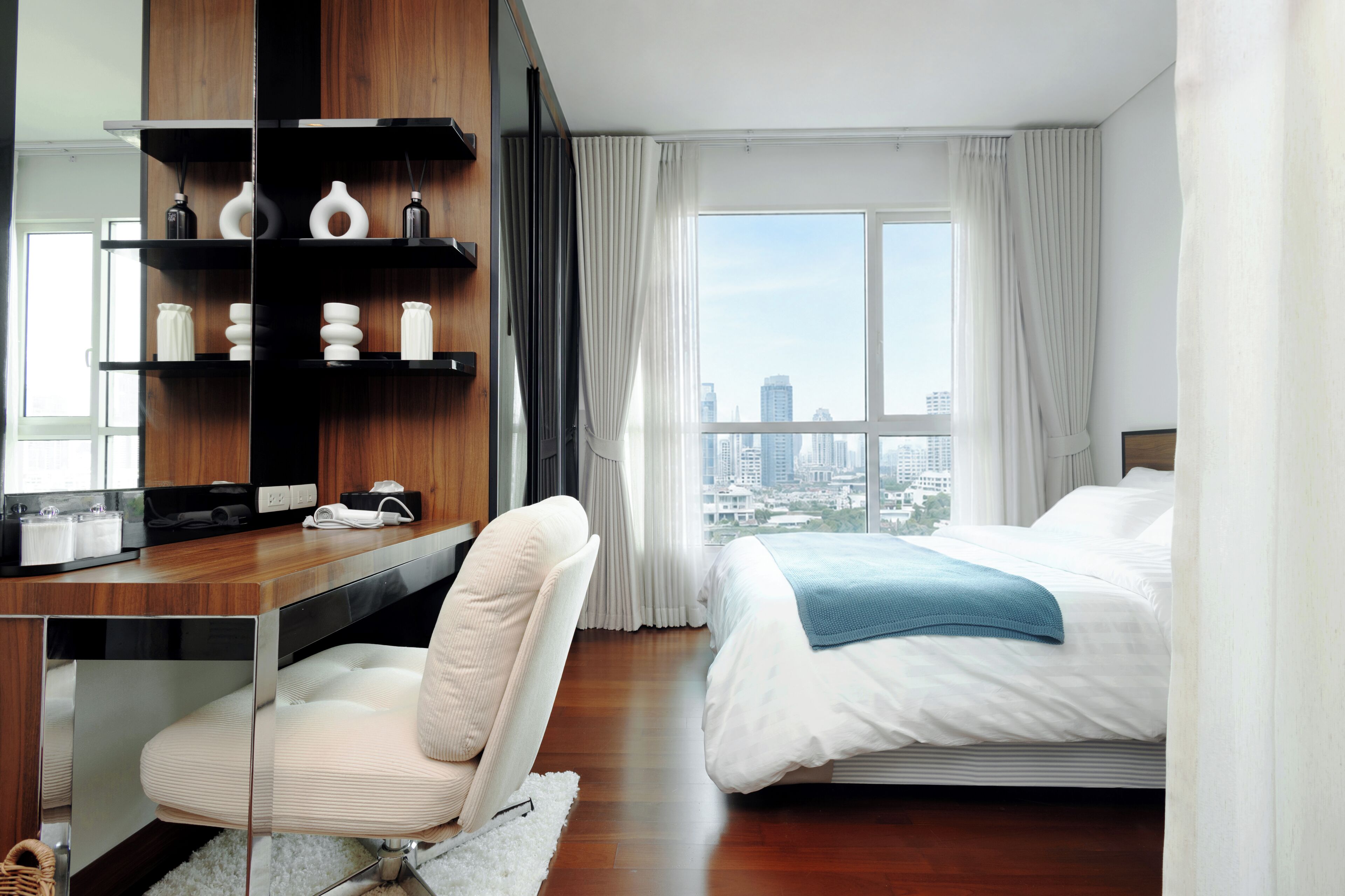 A hotel room with a bed, a desk with a chair, a large window with curtains, and a city view.