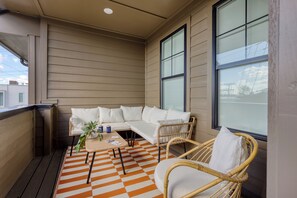 Ein überdachter Balkon mit einer beigen Couch, Korbstühlen und einem karierten Teppich.