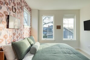 Ein Schlafzimmer mit einem Bett, grünen und weißen Kissen, floralem Wandpapier und zwei Fenstern.