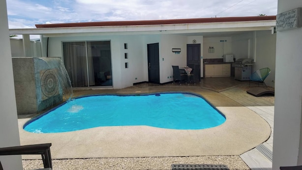 A swimming pool with a curved edge, a covered outdoor area with a kitchen, and a sign with a message. - Acogedora casa de playa, Condominio Malaga Bejuco (Bejuco)
