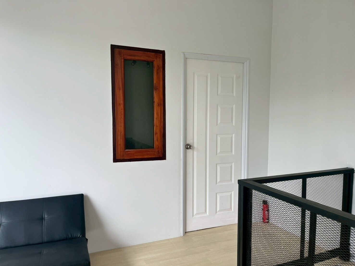 A room with a white door, a wooden-framed window, a black leather sofa, and a black metal railing.