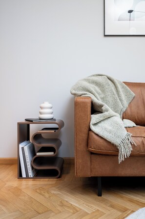 Un sofá de cuero marrón con una manta, una mesita de madera con libros y un jarrón blanco, y un cuadro en la pared.