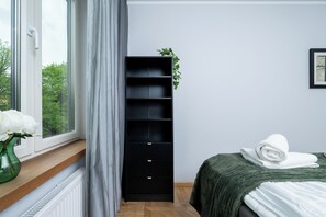 Un dormitorio con cama, ventana con cortinas, radiador, estantería negra y un cuadro en la pared.