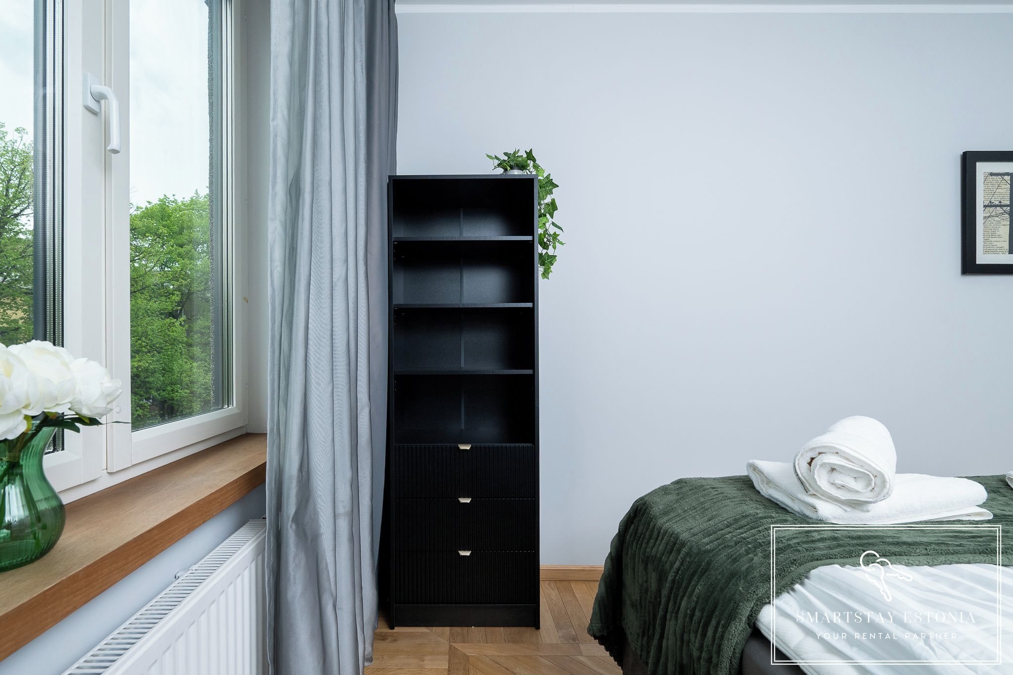 A bedroom with a bed, a black storage unit, a window with curtains, and a green plant on top of the unit.
