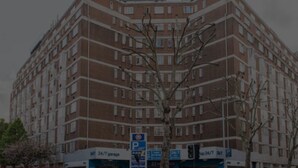 Un edificio de varios pisos con fachada de ladrillo, múltiples ventanas y un letrero de estacionamiento que indica disponibilidad las 24 horas.