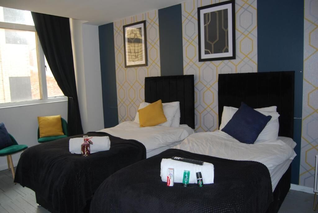 A hotel room with two beds, each with a black headboard and white bedding. There are pillows on the beds, and a small table with toiletries. The room has a window with curtains, and the walls have a geometric pattern.