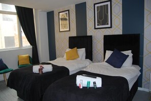 A hotel room with two beds, each with a black headboard and white bedding. There are pillows on the beds, and a small table with toiletries. The room has a window with curtains, and the walls have a geometric pattern. - Leicester Central Stay Near LRI and Uni (Leicester)