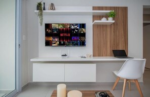 Un salón moderno con un televisor de pantalla plana montado en una pared blanca, una computadora portátil sobre un estante de madera, una silla blanca y una mesita con una vela.