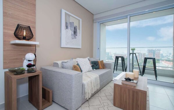 Una sala de estar moderna con un sofá gris, una mesa de centro, una mesita de noche con una tetera, una lámpara y vistas a la ciudad a través de amplios ventanales.