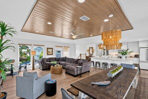 A modern living space with a wooden ceiling, a grey sectional sofa, a dining table with a plant centerpiece, and a kitchen area with white cabinets. - Waterfront Canal Home w/ Private Dock |Sleeps 11 (Rockport)