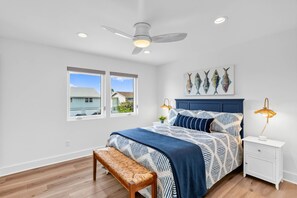 Quarto com cama, mesas de cabeceira, um banco, ventilador de teto e janela com vista para casas.