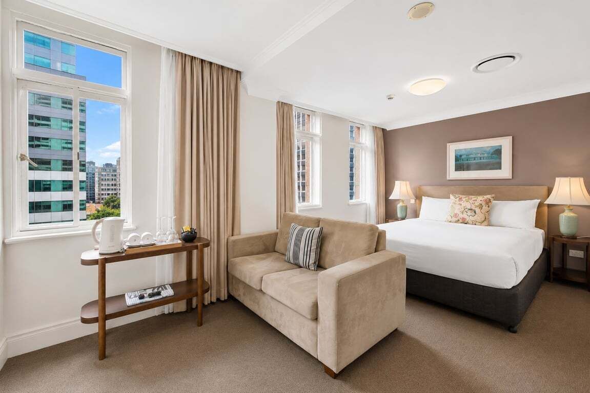 A hotel room with a bed, a sofa, a side table with a coffee maker, and a window with curtains.
