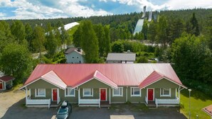 A row of single-story buildings with red roofs, a parking area, and a forested hillside in the background. - Apartment A 3 Talja, Private Sauna By Lapland Host (Rovaniemi)