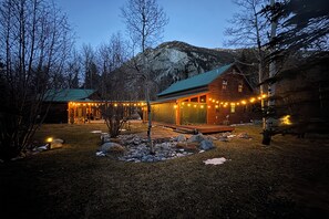 Une cabane en rondins surmontée d’un toit végétalisé, entourée d’un sentier en pierre et illuminée de guirlandes lumineuses.