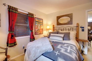 Une chambre à coucher avec un lit, une table de chevet, une lampe, une fenêtre avec des rideaux, une affiche accrochée au mur et un miroir.
