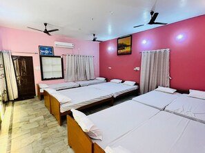 A hotel room with three single beds, a ceiling fan, an air conditioning unit, a window with curtains, and a painting on the wall.