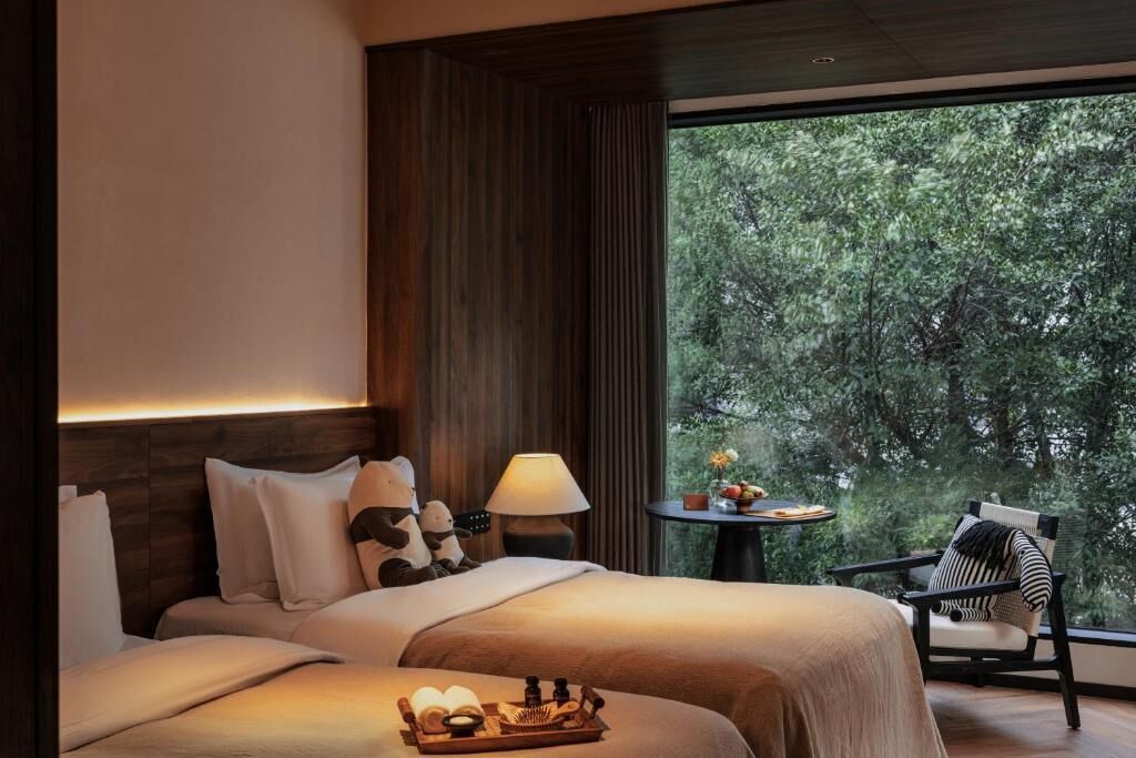 A hotel room with two beds, a nightstand with a lamp, a small table with fruit, and a view of trees through large windows.