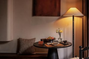Une table ronde avec une lampe, un saladier de fruits, un vase de fleurs et un coussin.