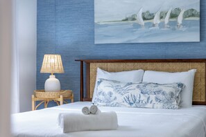 Ein ordentlich bezogenes Bett mit weißer Decke und zwei Kissen, ein Korbnachtkästchen mit einer Lampe und ein Bild von Segelbooten an der Wand.