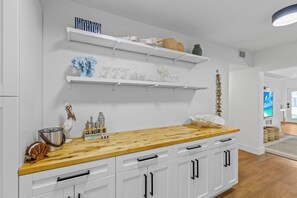 A kitchen with white cabinetry, a wooden countertop, and shelves with glassware and decorative items. - Private Pool + Firepit + Beach Gear | Short Drive to Beach + Las Olas | Al Mare (Fort Lauderdale)