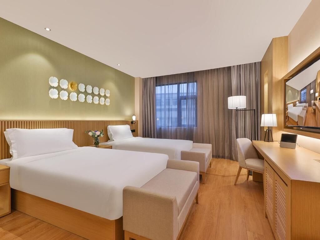 Habitación de hotel con dos camas, un escritorio y una silla. Cuenta con un ventanal con cortinas, una lámpara de pared y un adorno en la pared.