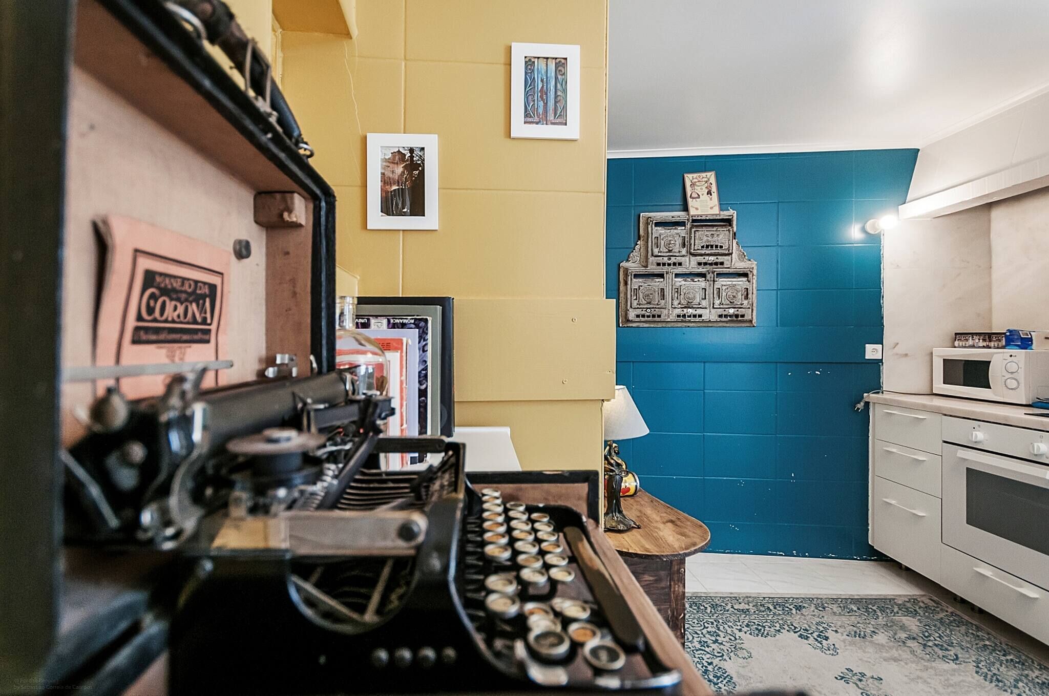 Una máquina de escribir antigua, un cuadro enmarco, una pared de azulejos azules y un microondas en una zona de cocina.