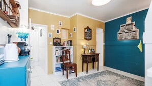 Un pasillo con un armario azul, una estantería, una silla, una mesita con una lámpara y un muro con cuadros enmarcados.