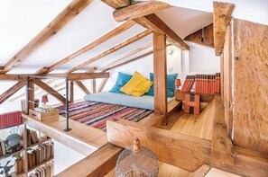 Quarto estilo loft com vigas de madeira aparentes, um sofá azul com almofadas coloridas, um tapete estampado e uma pequena mesa de madeira com um globo e livros.
