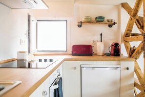 Eine kleine Küche mit Fenster, einem Holzbodenregal mit Schüsseln, einem roten Koffer und einem weißen Geschirrspüler.