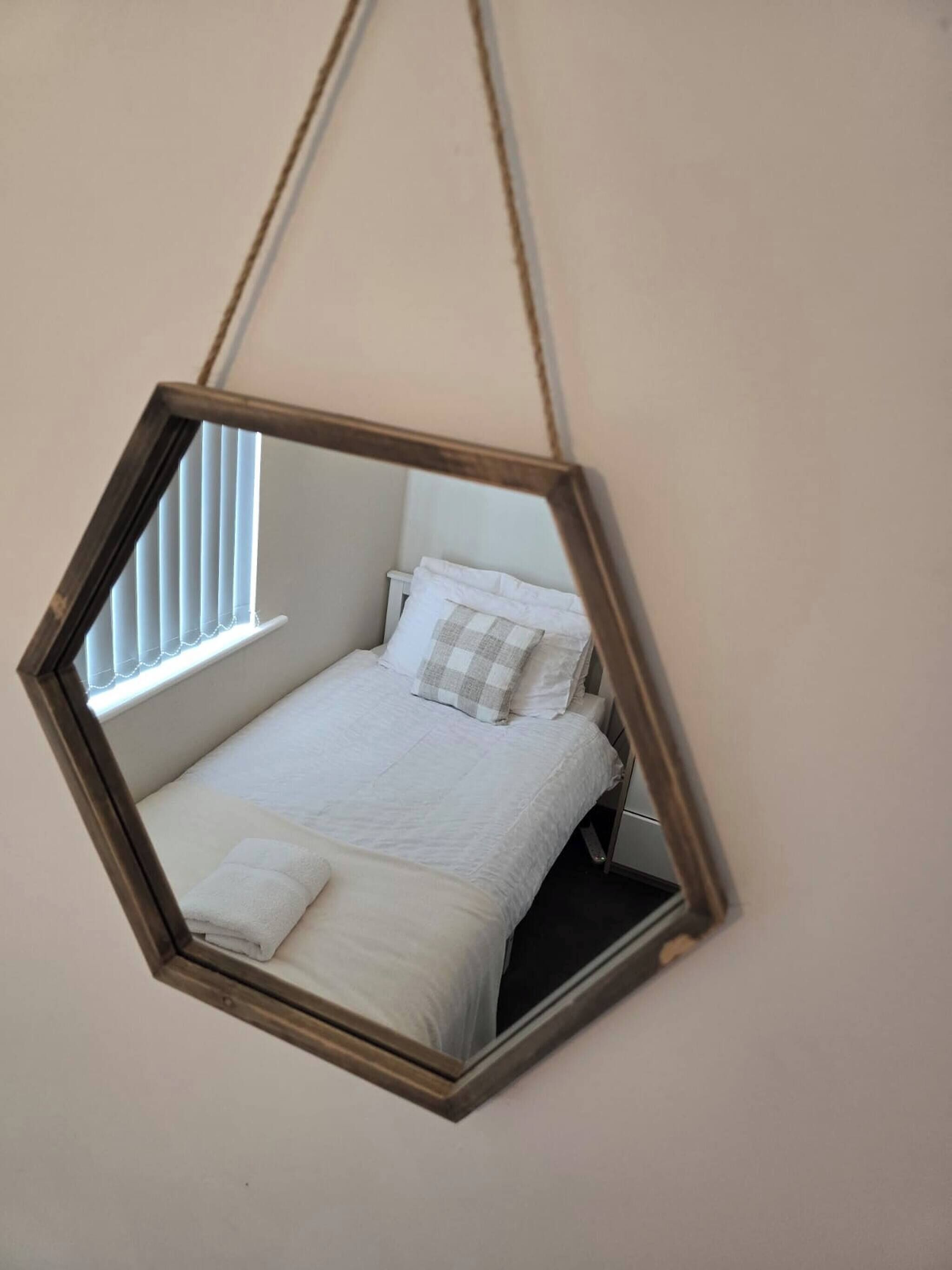 A hexagonal mirror hanging in a room with a bed, pillows, and a window.