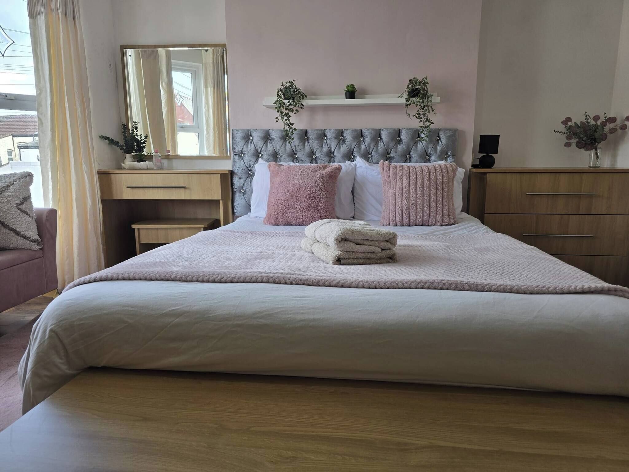 A neatly made bed with a headboard, a bedside table, a mirror, and a shelf with plants.