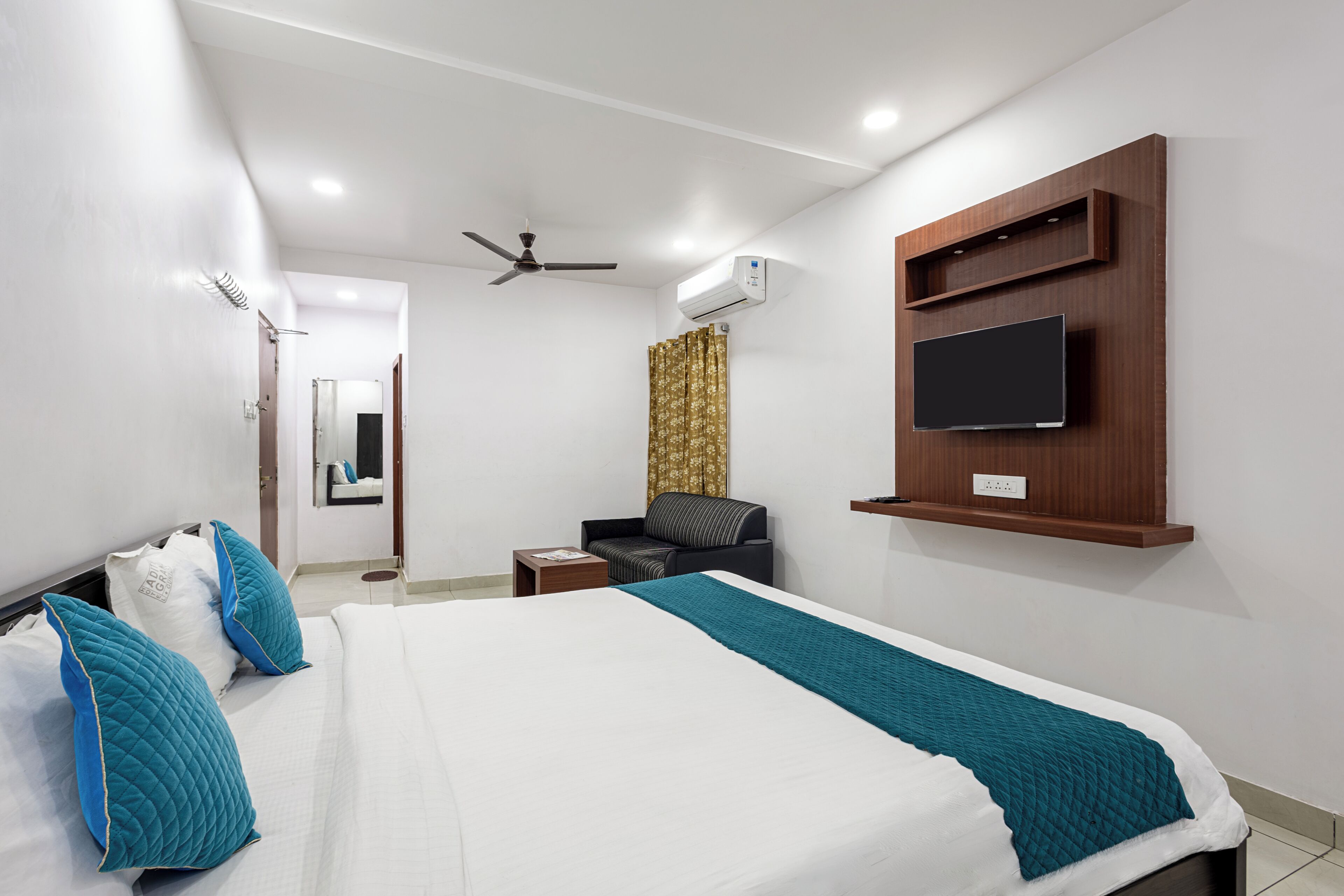 Una camera d'hotel con un letto, una TV montata a parete, una ventola a soffitto, un condizionatore e un tavolino con una sedia.