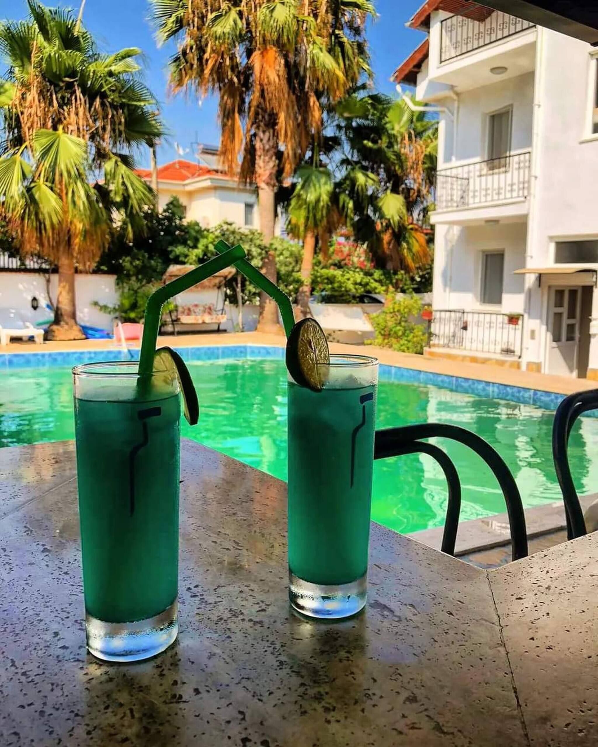 Två glas med grön dryck och sugrör står vid poolkanten, omgivna av palmer och en byggnad med balkonger.