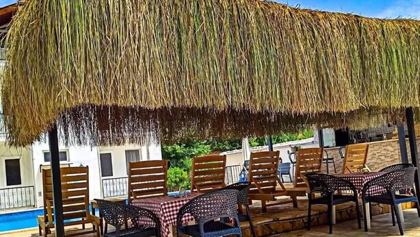 Un espacio exterior con mesas y sillas bajo un techo de paja, con vista a una piscina.