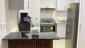 Et moderne køkken med en sort bordplade, to barstole, en kaffemaskine, en vanddispenser, en komfur og et køleskab, samt hvide skabe.