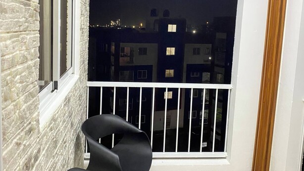 En balkon med udsigt til bygninger om natten, en sort stol og et hvidt gelænder.