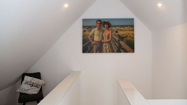 A painting of a couple on a beach, a white wall, and a small black chair with a pillow. - Lüttje Sünn - Lüttje Sünn - Your oasis of well-being in Norddeich (Norden)