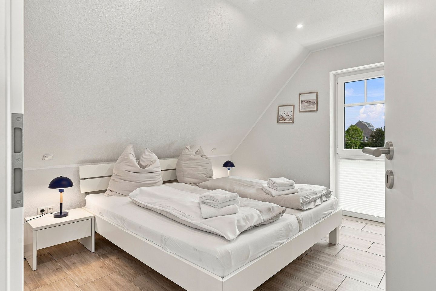 Een moderne slaapkamer met een schuine kap, een bed met witte lakens, twee nachtkastjes met lampen en een raam met uitzicht.