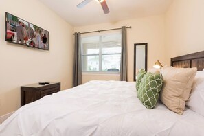 Een slaapkamer met een bed, een nachtkastje, een plafondventilator, een raam met gordijnen, en een schilderij aan de muur.