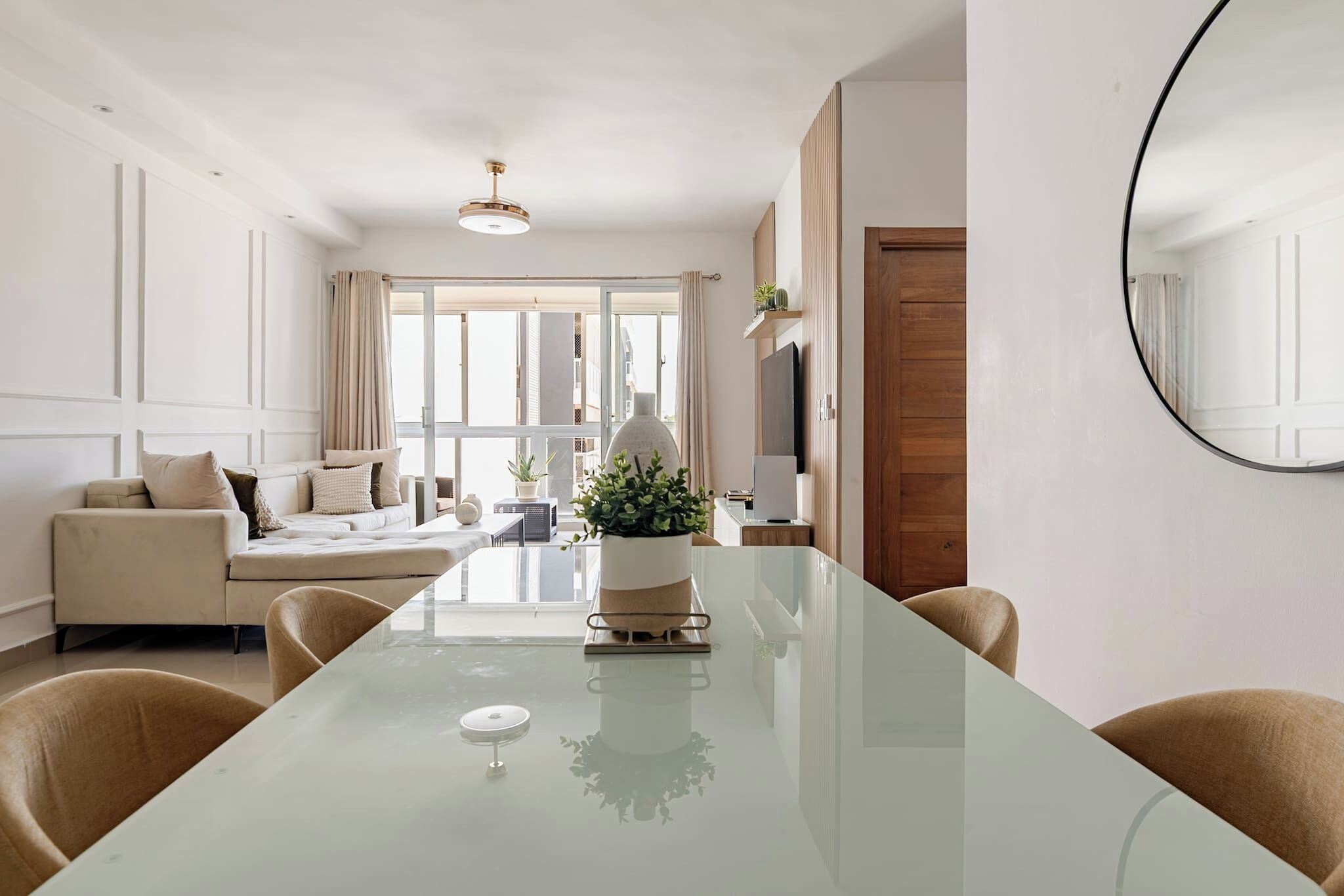 Una sala moderna con una mesa de comedor de vidrio, sofás color beige, una planta en maceta y un espejo grande en la pared.