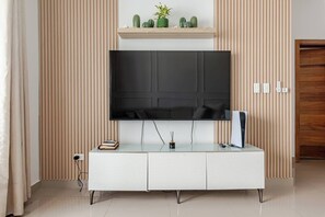 Ein modernes Wohnzimmer mit einem an der Wand montierten Flachbildfernseher, einer weißen Unterhaltungsecke mit Glastisch und einer Holztür im Hintergrund.